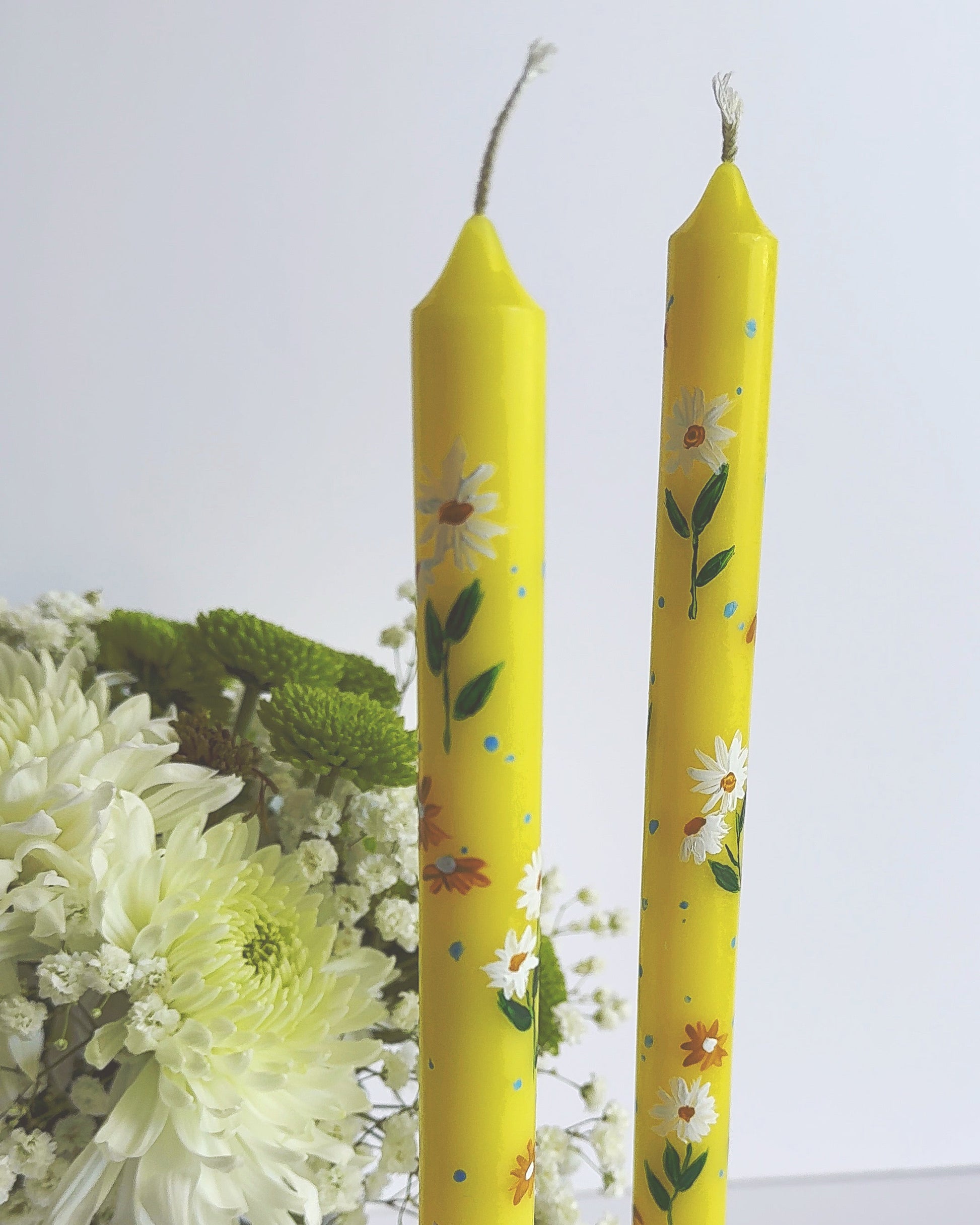 Two yellow candles with floral patterns on a white surface with flowers in the background