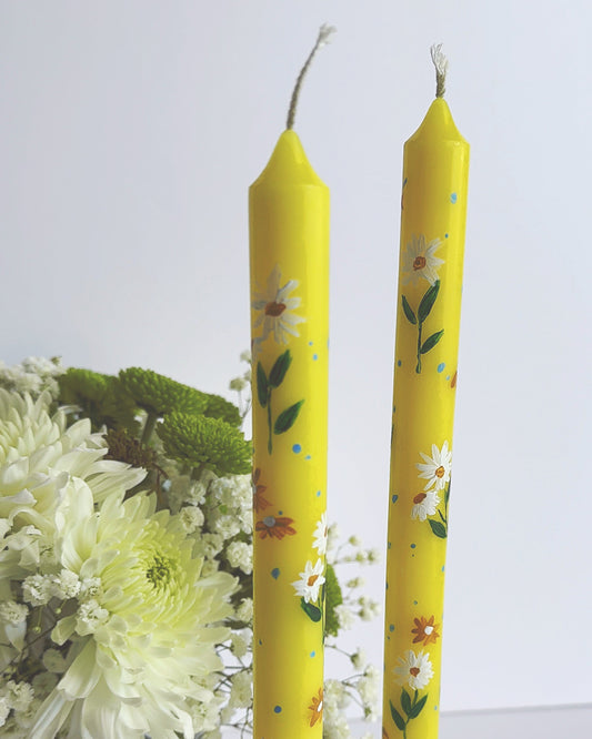 Two yellow candles with floral patterns on a white surface with flowers in the background
