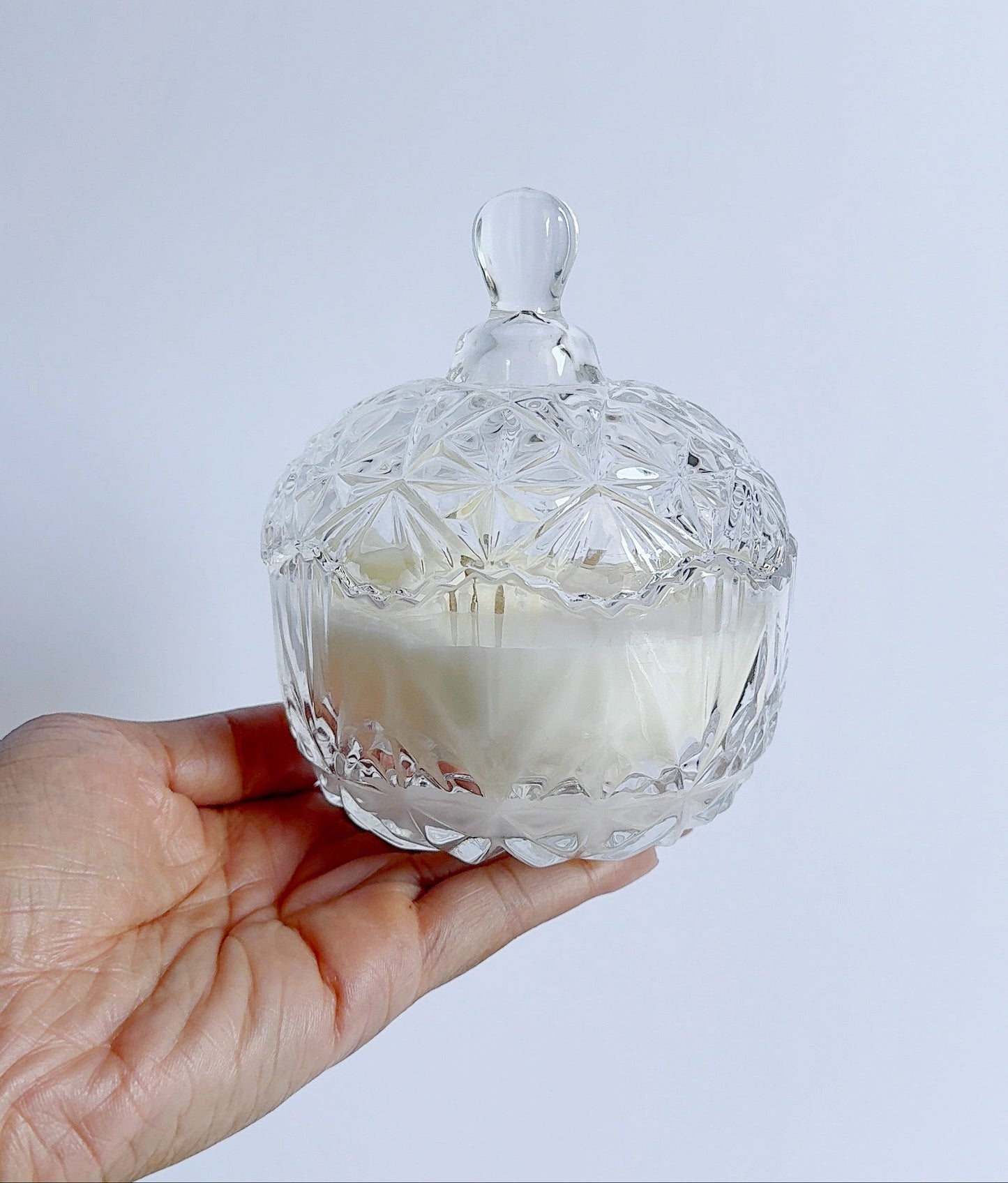Hand holding a clear glass candle dish with a white candle inside against a light gray background