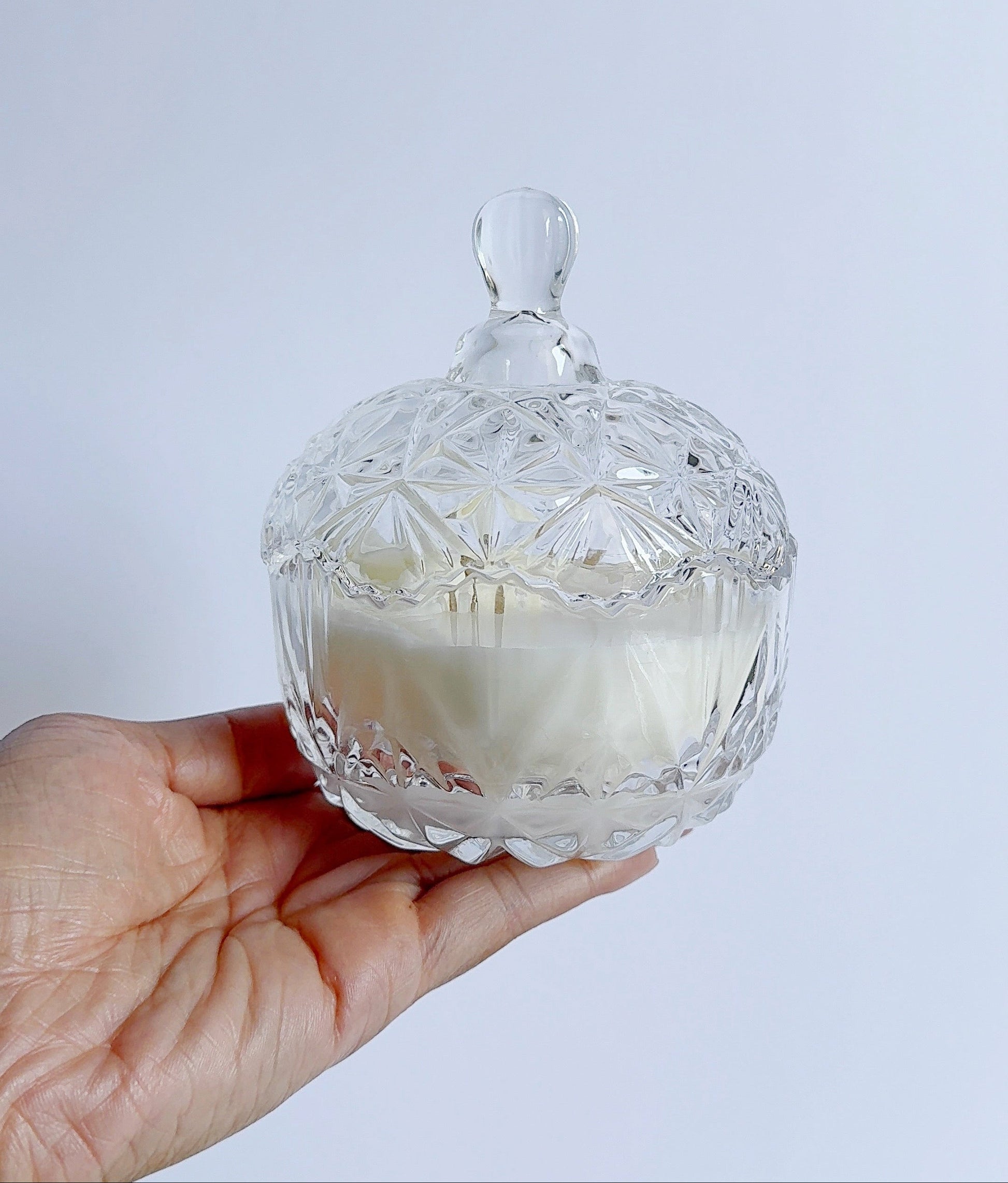 Hand holding a clear glass candle dish with a white candle inside against a light gray background