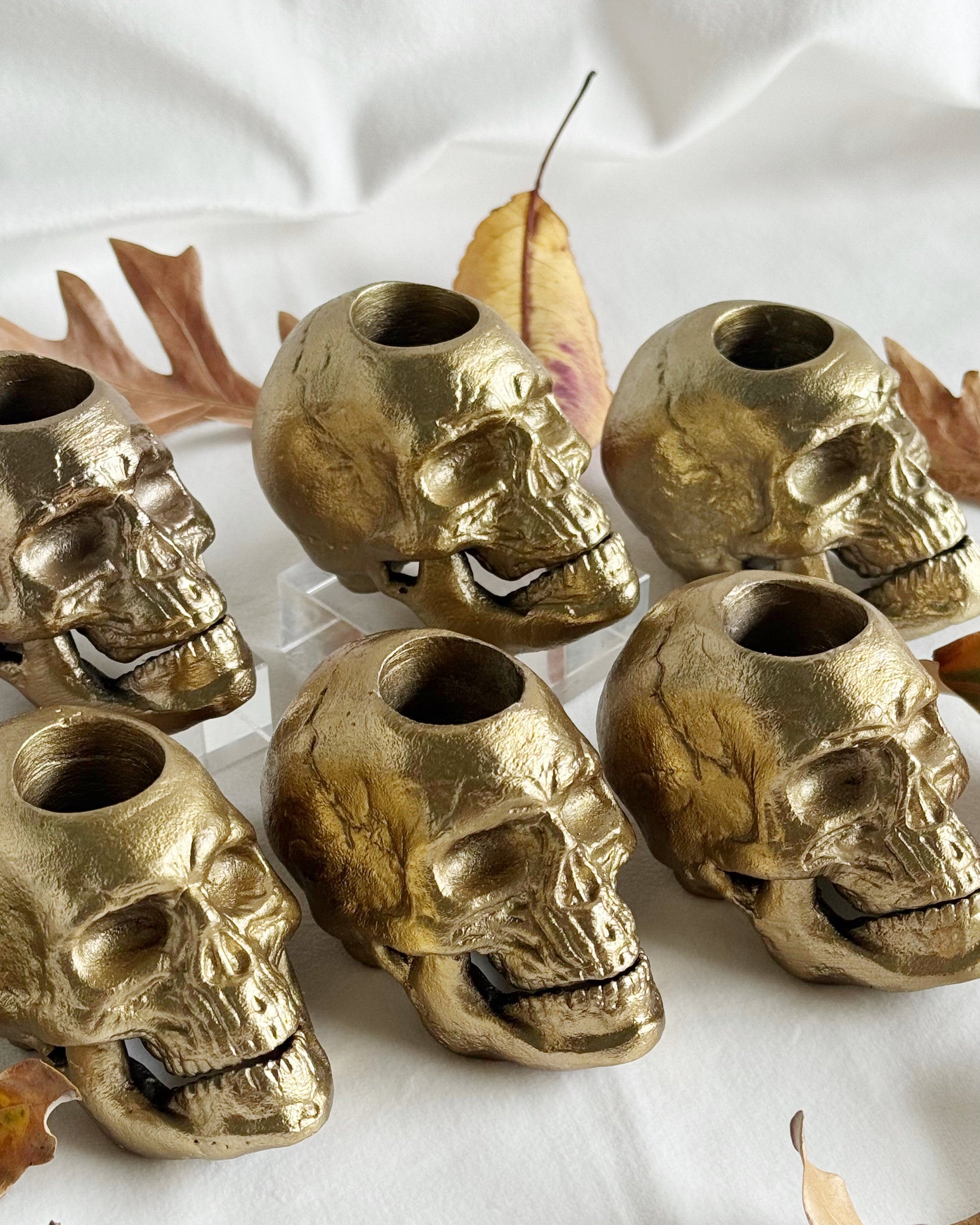 Gold skull-shaped candle holders on a white surface with autumn leaves.