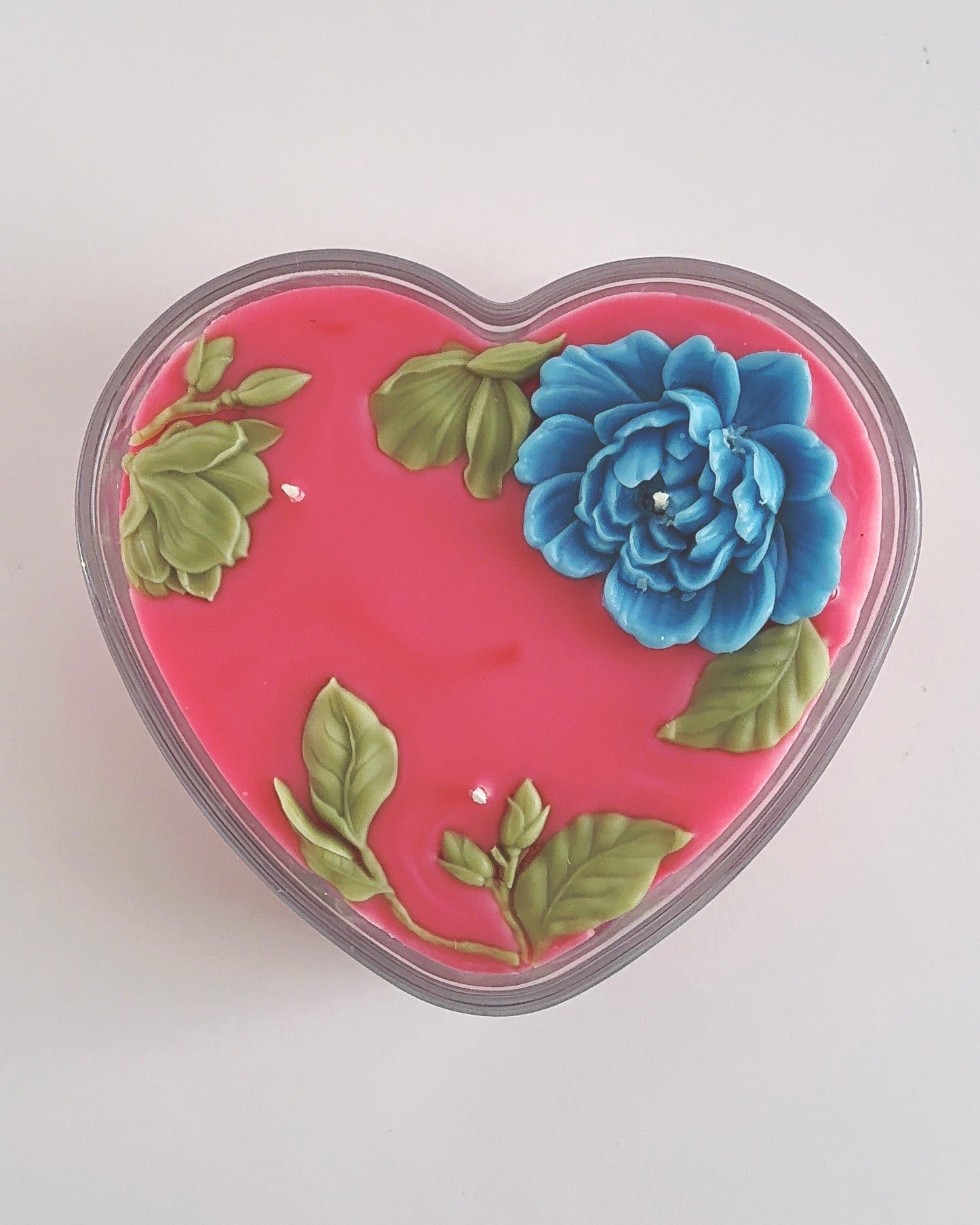 Heart-shaped object with a blue flower and green leaves on a light gray background